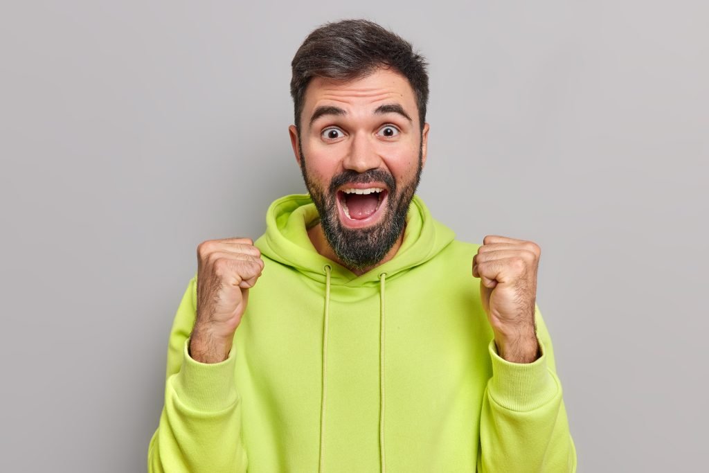 Cheerful bearded handsome man raises fists in victory gesture celebrates success or triumph exclaims from big joy wears casual green sweatshirt isolated over grey background. Yes I did it finally